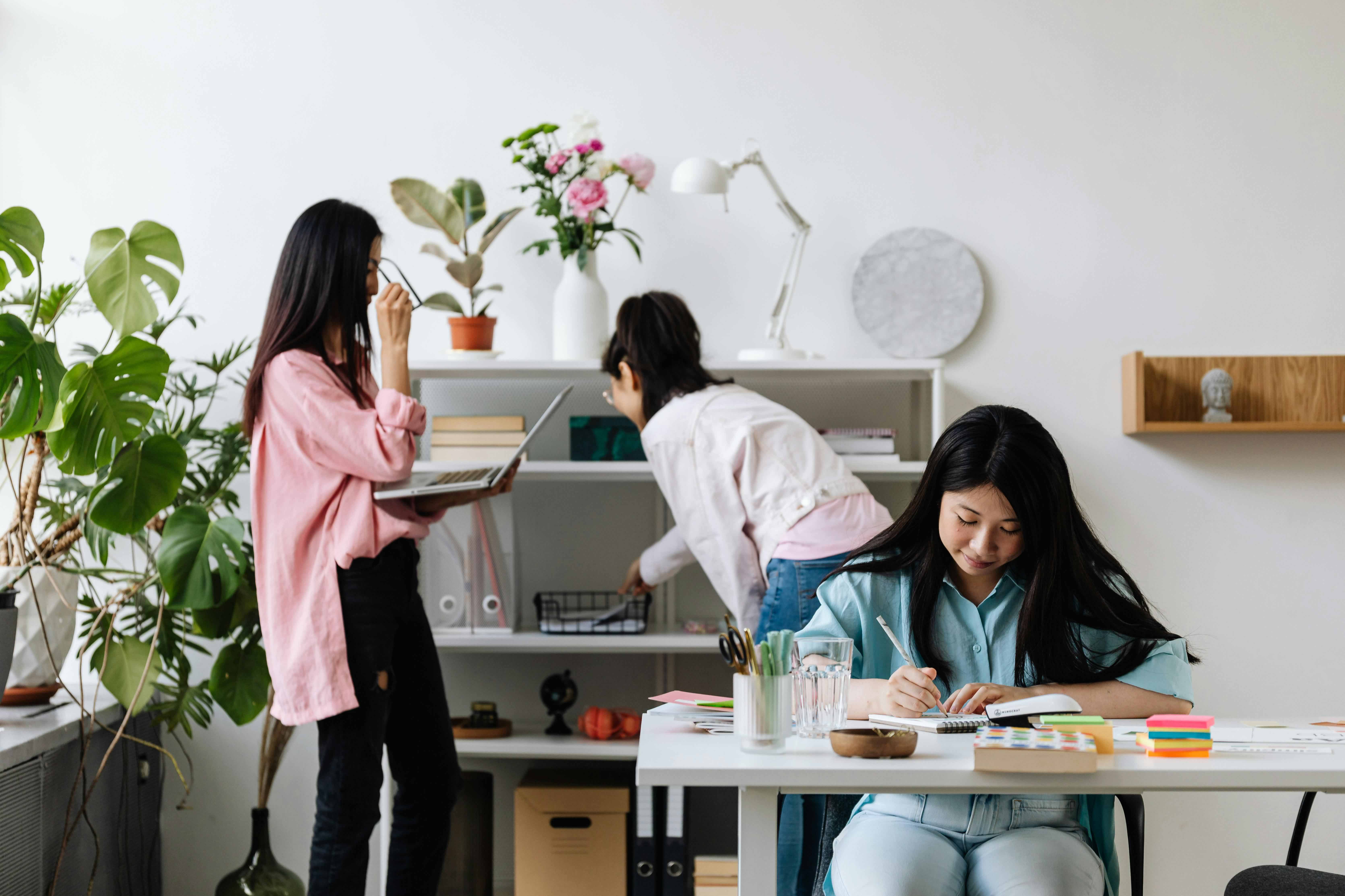 Three women: one taking notes, another working on a laptop, and the third looking at a shelf
